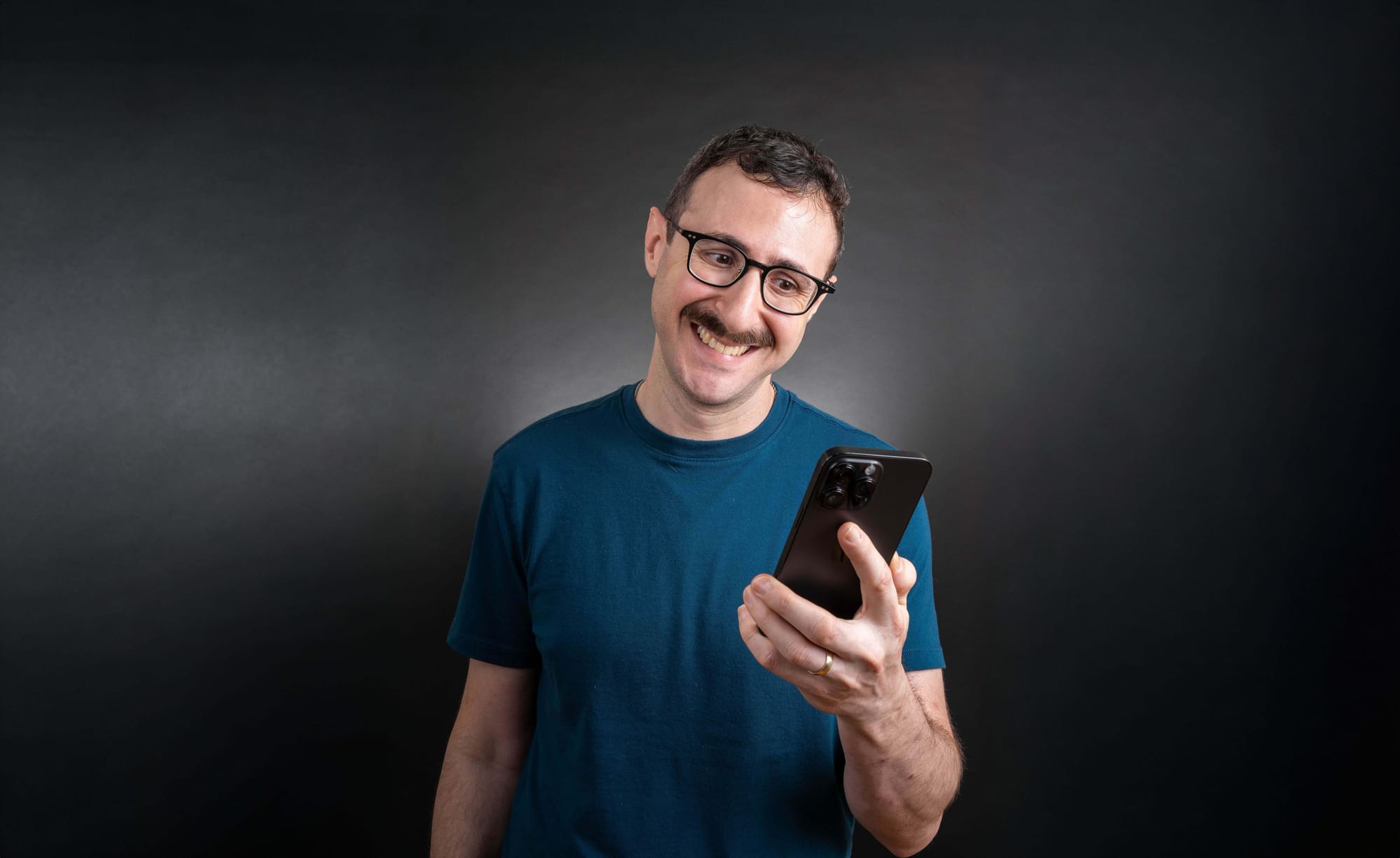Dann Berg, a man with short hair, glasses, and a mustache looks down at a smartphone he’s holding in one hand. He’s wearing a teal T-shirt and standing against a dark, neutral background.